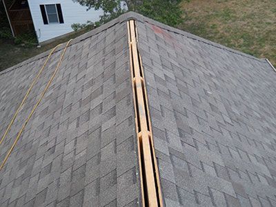 An unfinished roof ridge with an exposed wooden vent channel surrounded by grey asphalt shingles.
