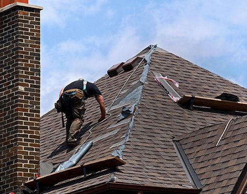 A person wearing a tool belt and safety harness works on the brown shingled roof of a house with a brick chimney.