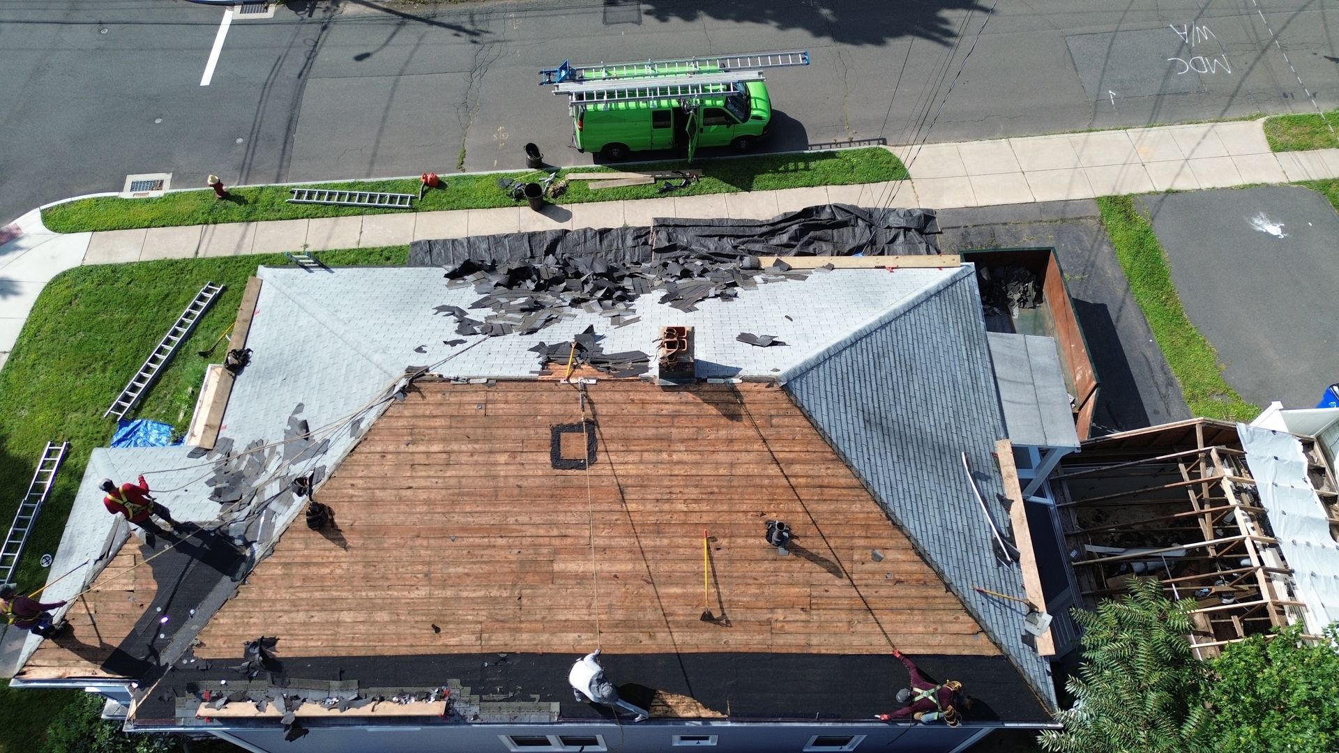 An aerial view shows workers performing a roof replacement on a house, with exposed wood decking and a nearby green van.