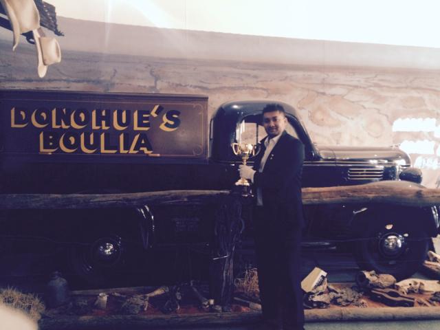 Man in suit holds a trophy, posing in front of a vintage truck and sign reading 