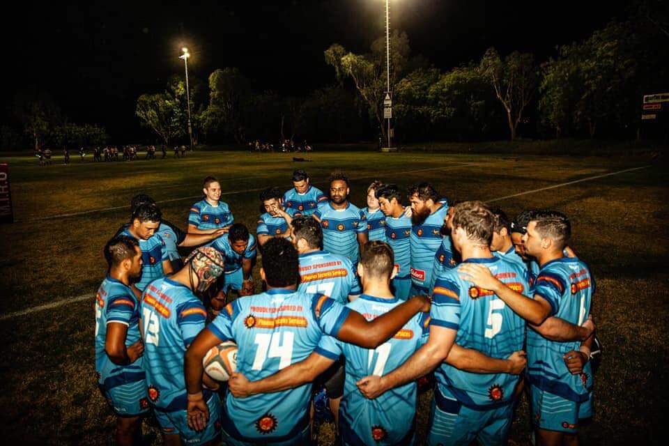 A rugby team in blue jerseys huddles together on a grass field at night. The players are lit by overhead stadium lights.
