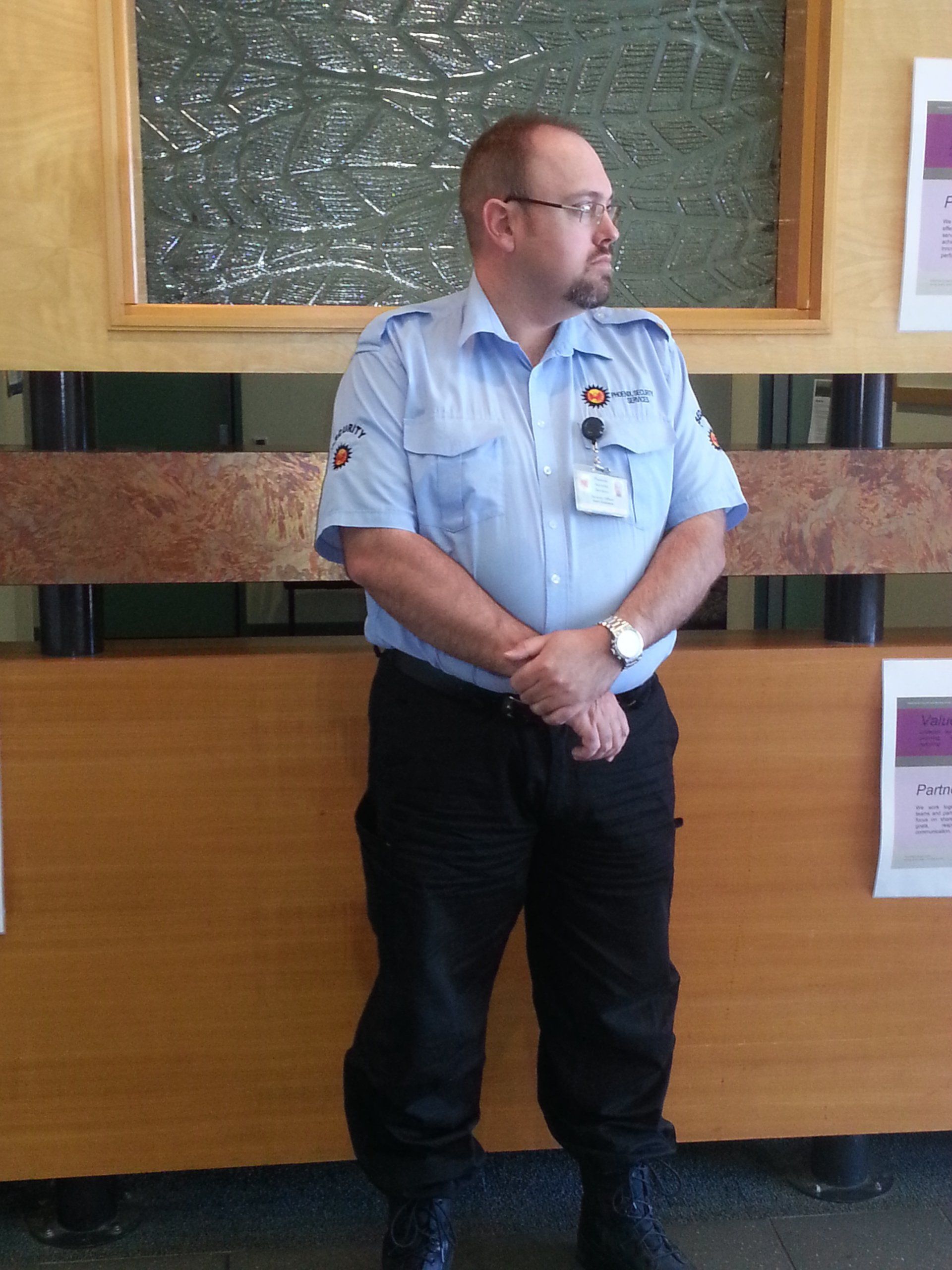 A man in a blue uniform stands, arms crossed, looking off to the side. He's in a room with a wooden wall.