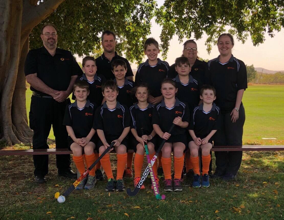 A youth hockey team in black and orange uniforms poses with their coaches on a sunny field.