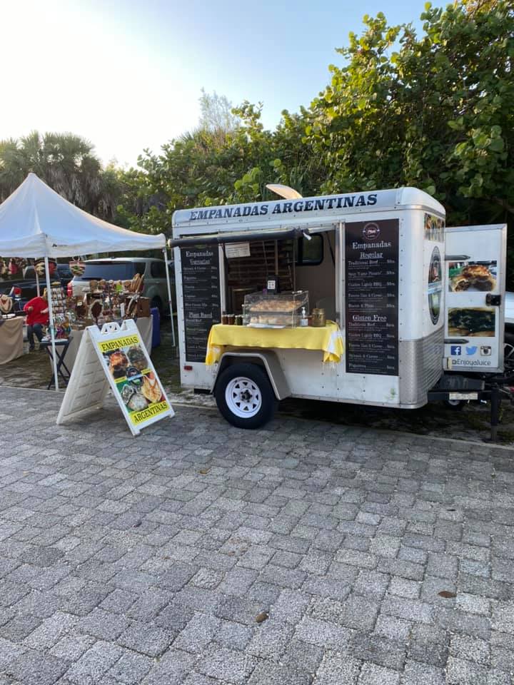 Food truck selling empanadas, with a menu board. White truck with a white tent and brick pavement.