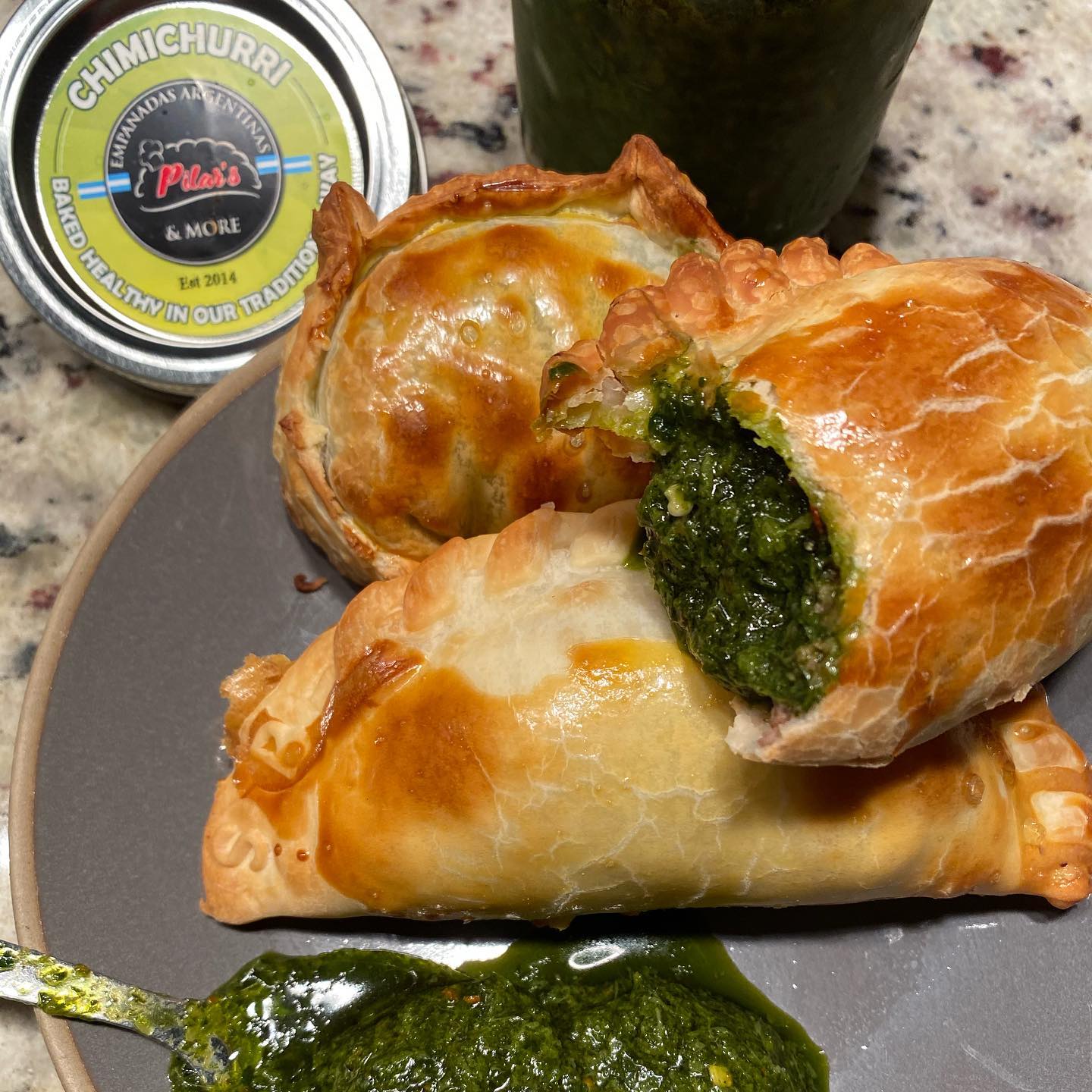 Two golden, baked empanadas, one cut open to reveal spinach filling, with chimichurri and an avocado.