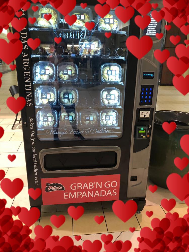 Vending machine for Grab 'N Go Empanadas, with several transparent compartments. Hearts frame the machine.