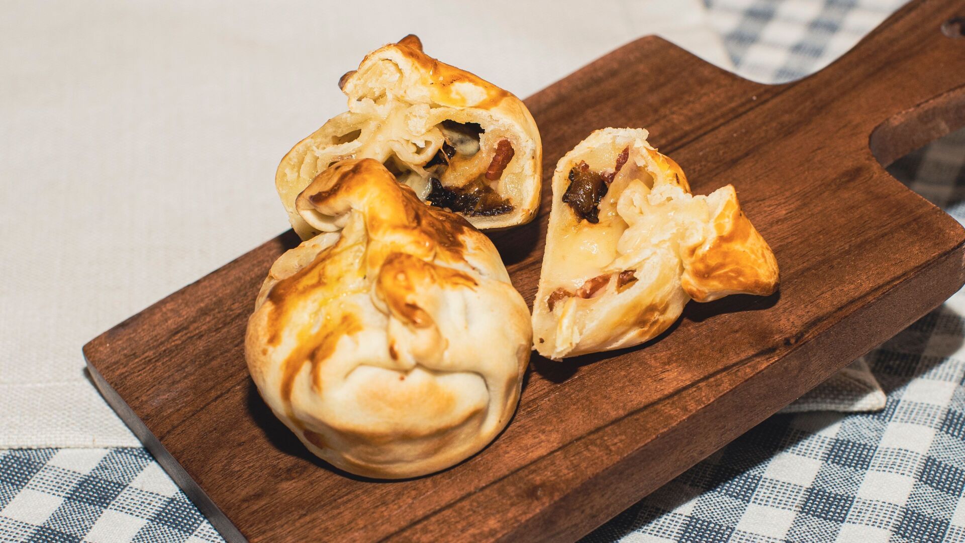 Three baked pastries on a wooden board, some split to reveal a dark filling, set on a checkered tablecloth.