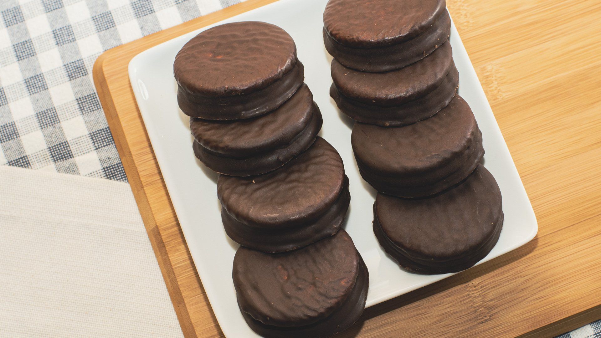 Stacks of chocolate alfajores on a white plate, on a wooden board, on a checkered cloth.