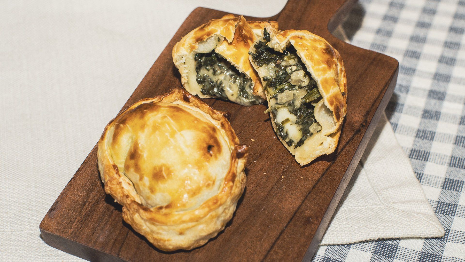 Two spinach and cheese pies on a wooden board; one is cut open, showing filling.