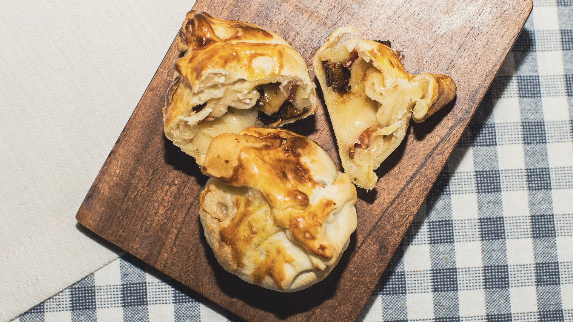 Empanadas on wooden board, golden crust, some cut open to show filling, on checkered tablecloth.