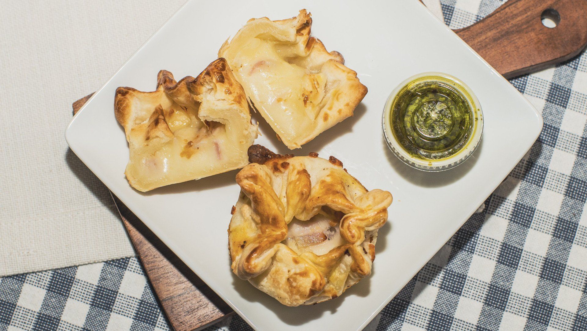 Three baked pastries on a white plate, with green dip. Plaid tablecloth and wooden cutting board in the background.