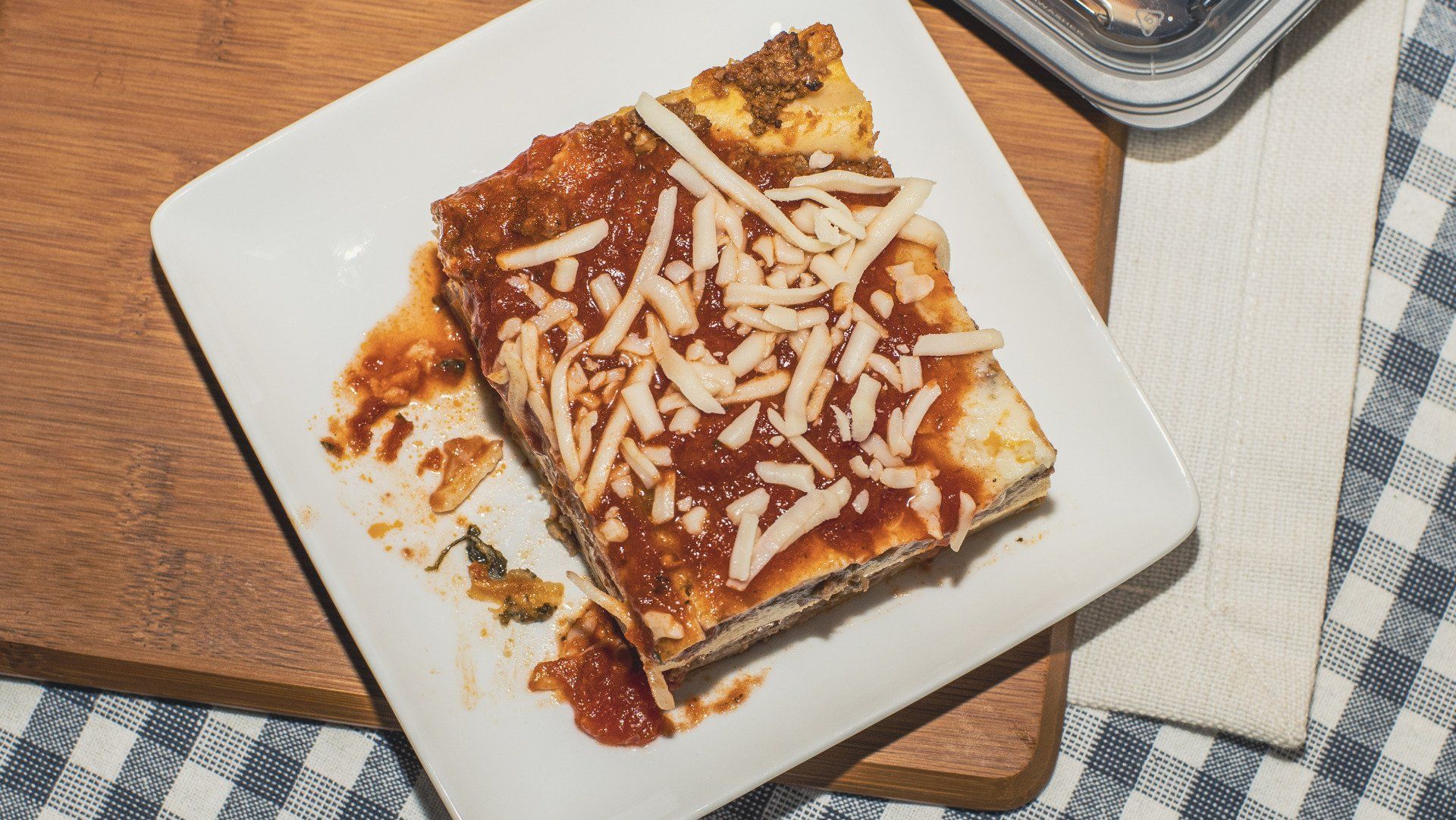 Square slice of lasagna on a white plate, topped with sauce and shredded cheese.