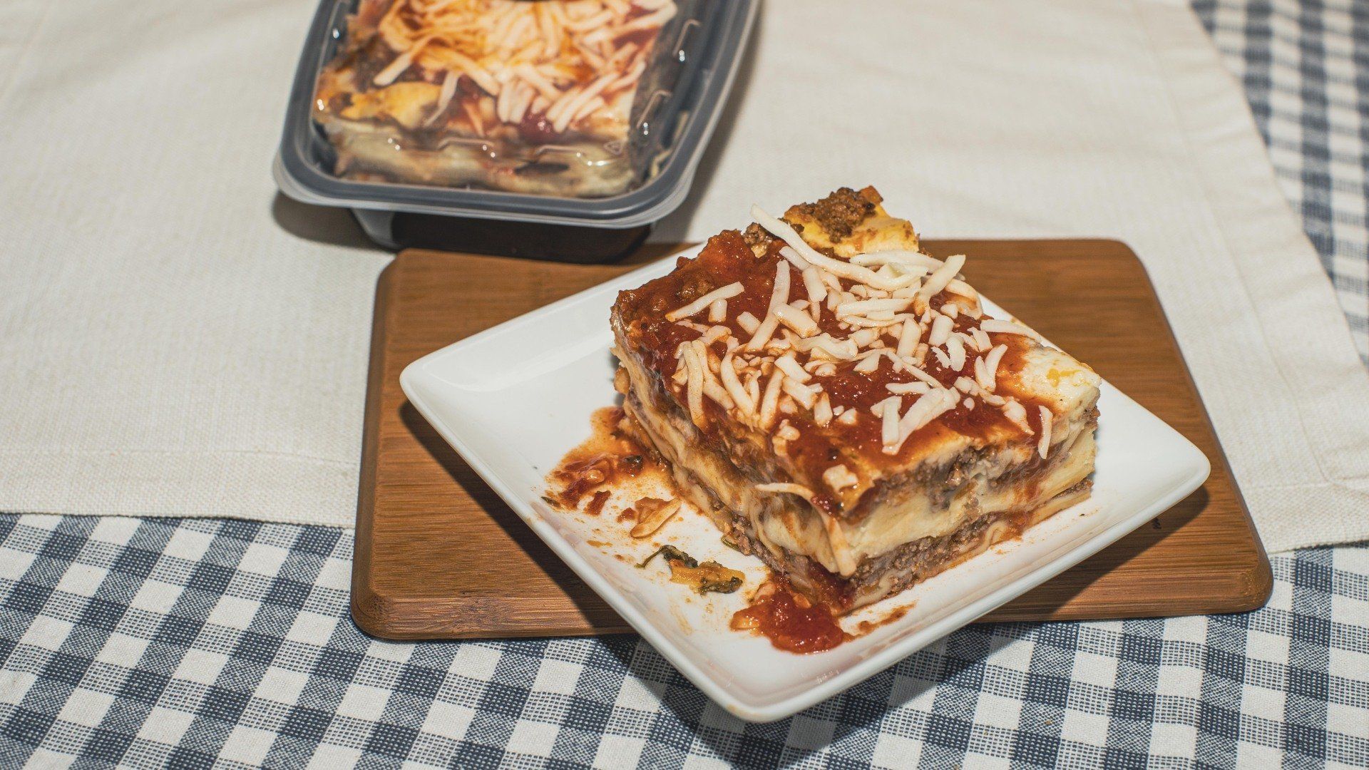 Lasagna on white plate and wooden board, with a container of lasagna in background on a checkered tablecloth.