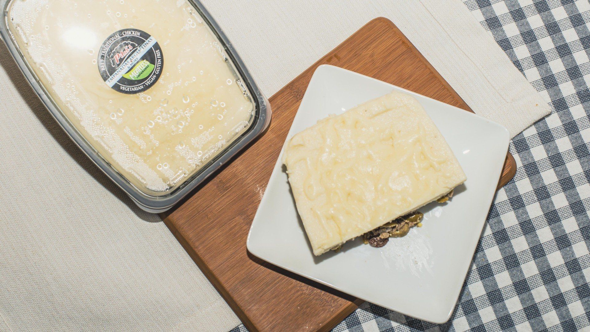 Overhead shot: a rectangular slice of food on a white plate, beside a container with the same food, and a wooden board.