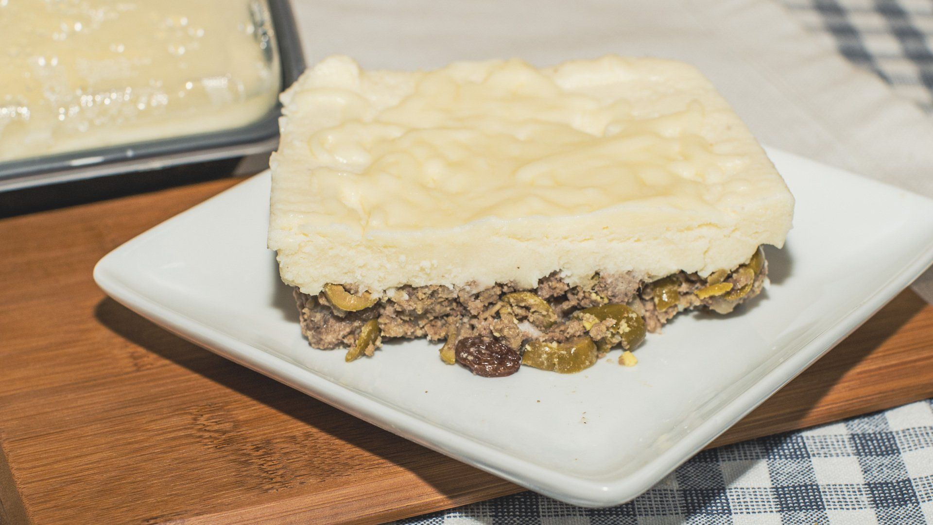 Square meat pie slice on a white plate atop a wooden board. Light crust tops a visible meat and veggie filling.