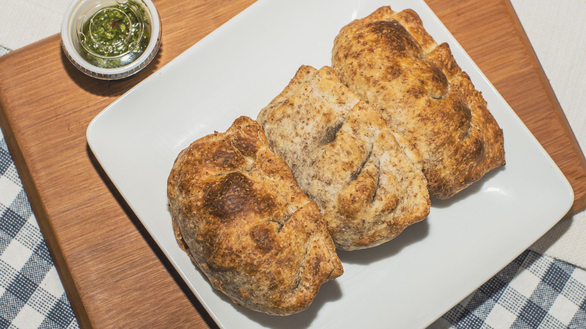 Three golden-brown biscuits on a white plate, served with a small bowl of green sauce on a wooden board.