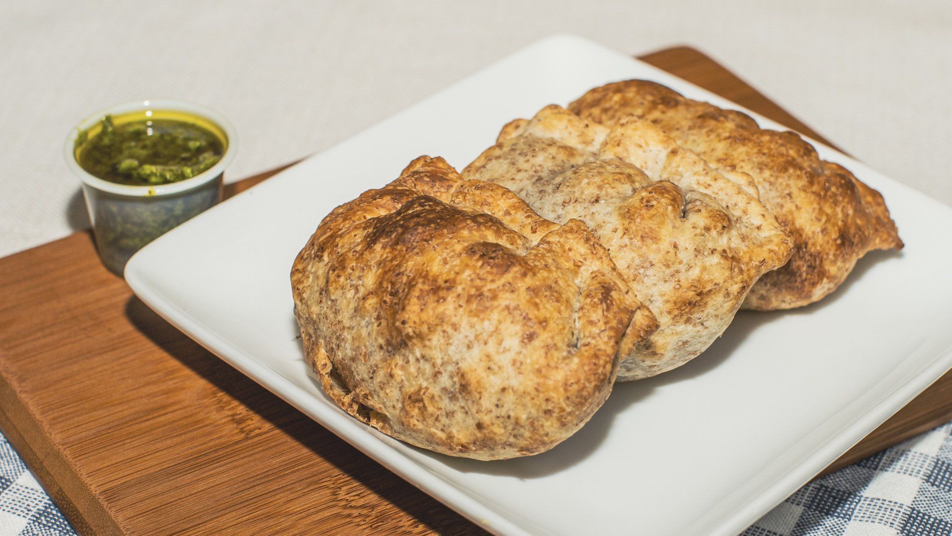 Three baked empanadas on a white plate with green dipping sauce, wooden serving board, and blue and white cloth.