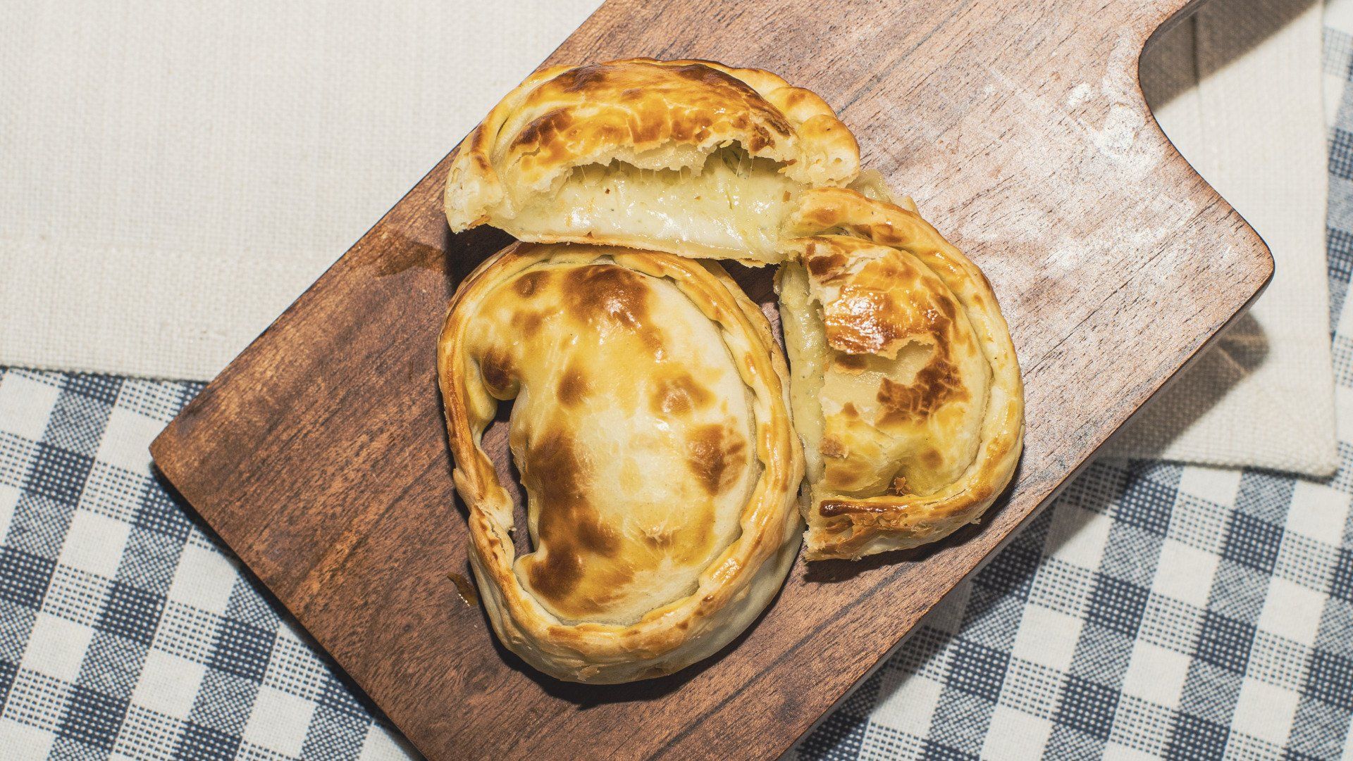 Empanadas on a wooden board, one cut open to show filling, on a blue checkered surface.