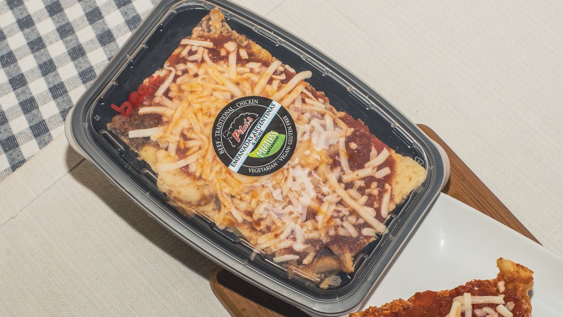 Prepared meal in a plastic container, topped with cheese and sauce, on a wooden board.