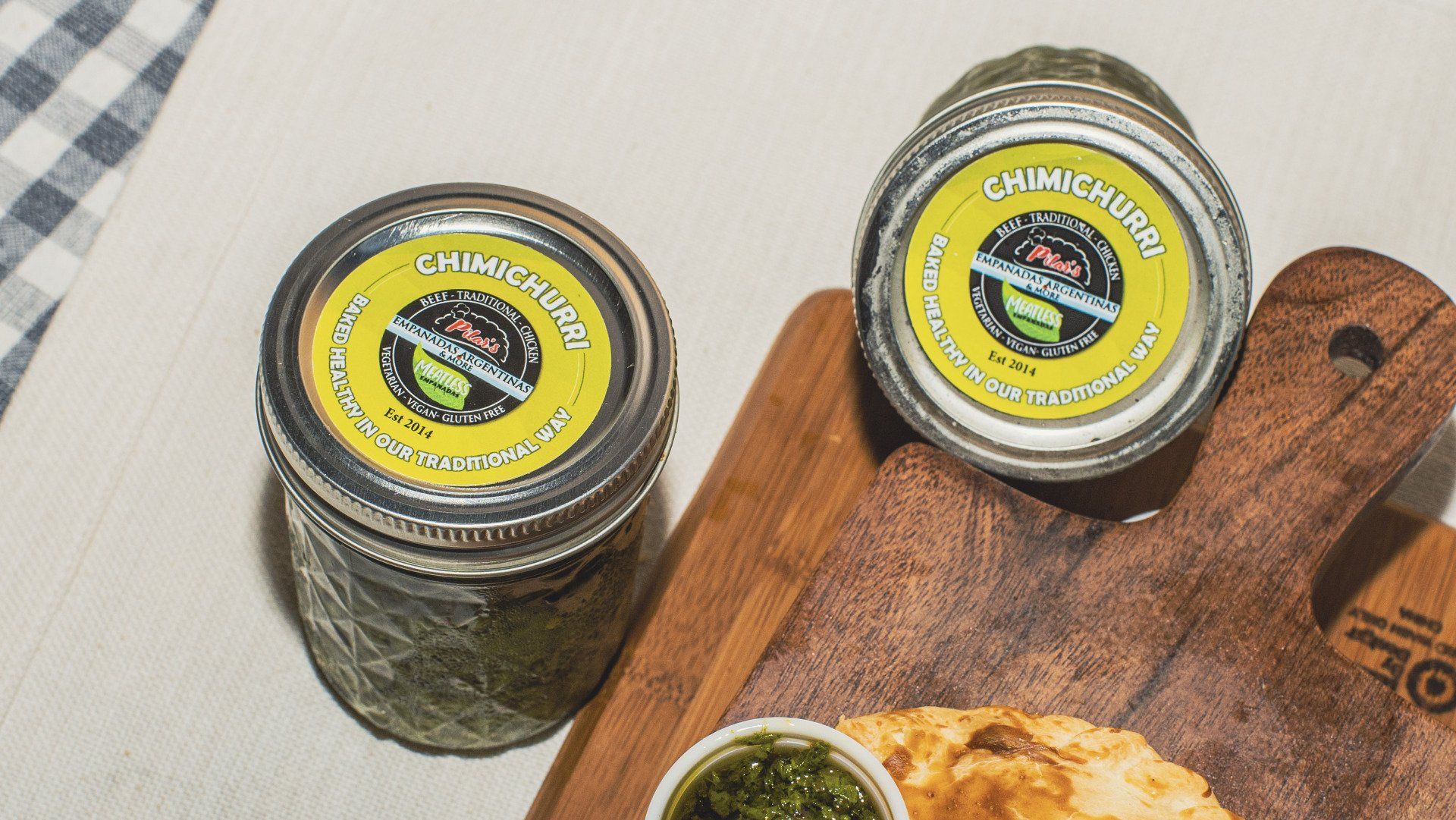 Two jars of chimichurri sauce on a wooden board, next to an empanada and small bowl of sauce.