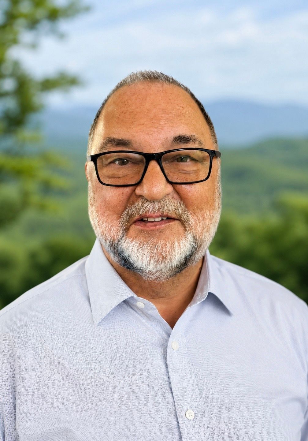 Man with gray beard, wearing a navy shirt, smiles outdoors with a green and yellow blurred background.