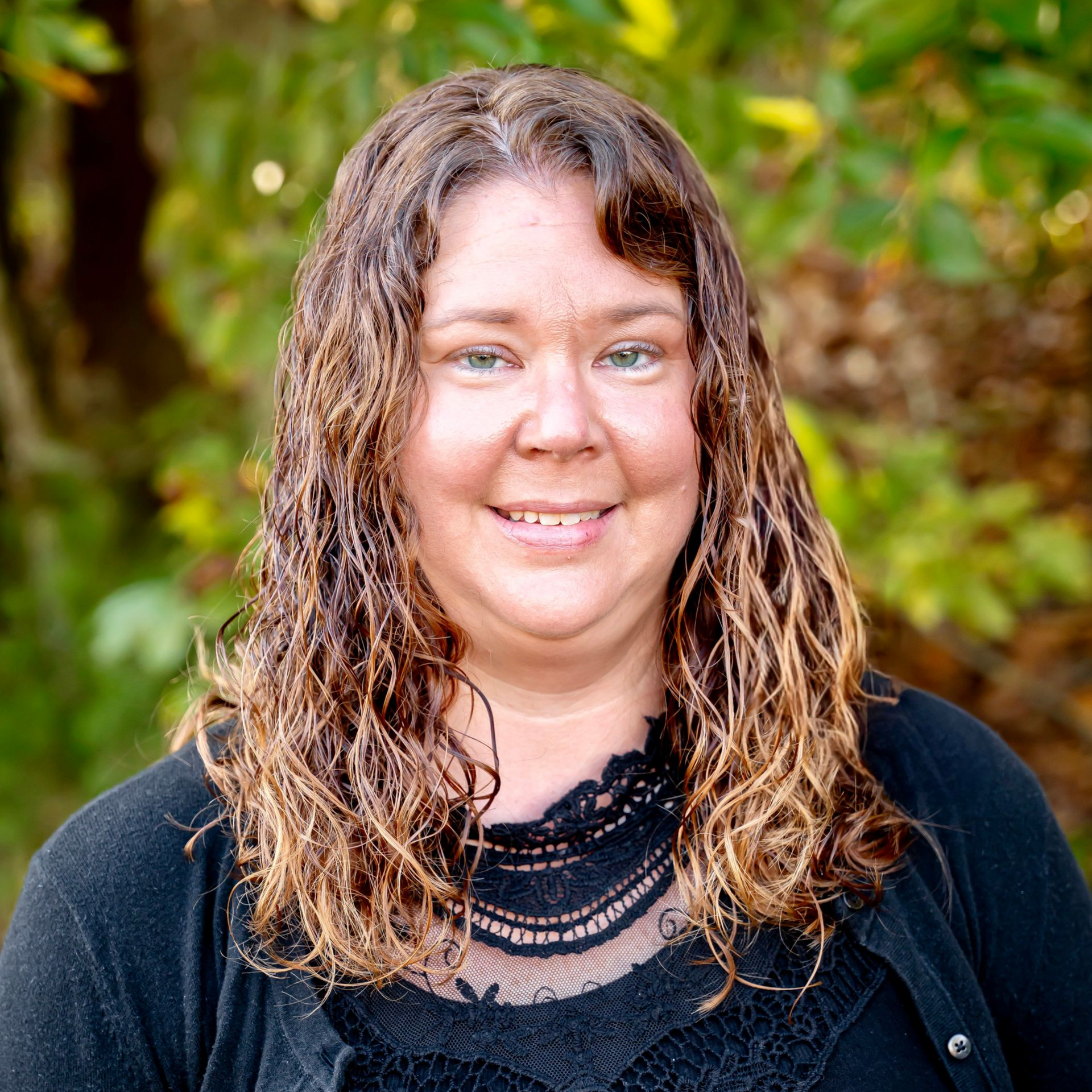 Woman with shoulder-length curly brown hair smiles, wearing a black cardigan, set against a blurred green background.