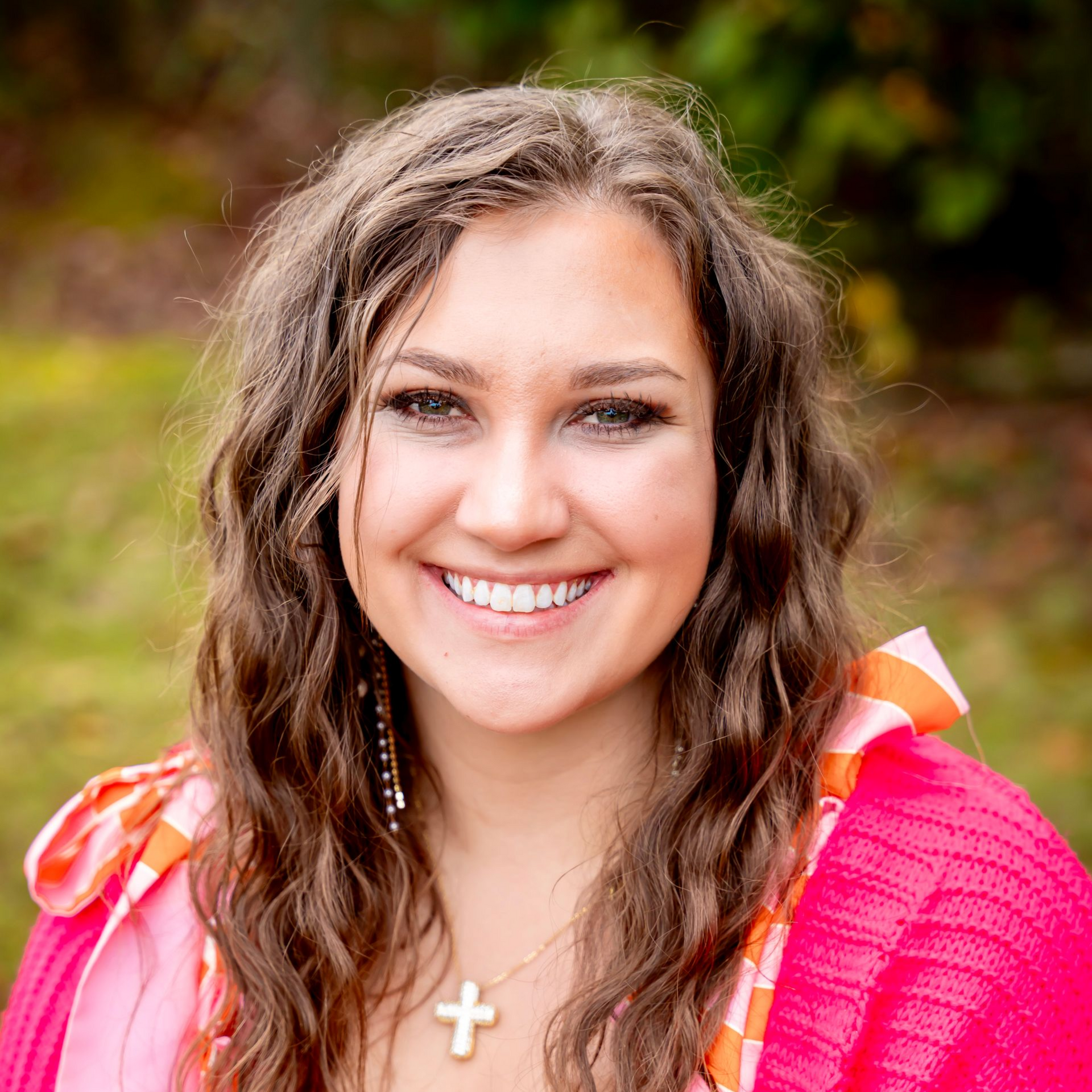 Woman smiling, wearing pink sweater over patterned top, gold cross necklace.