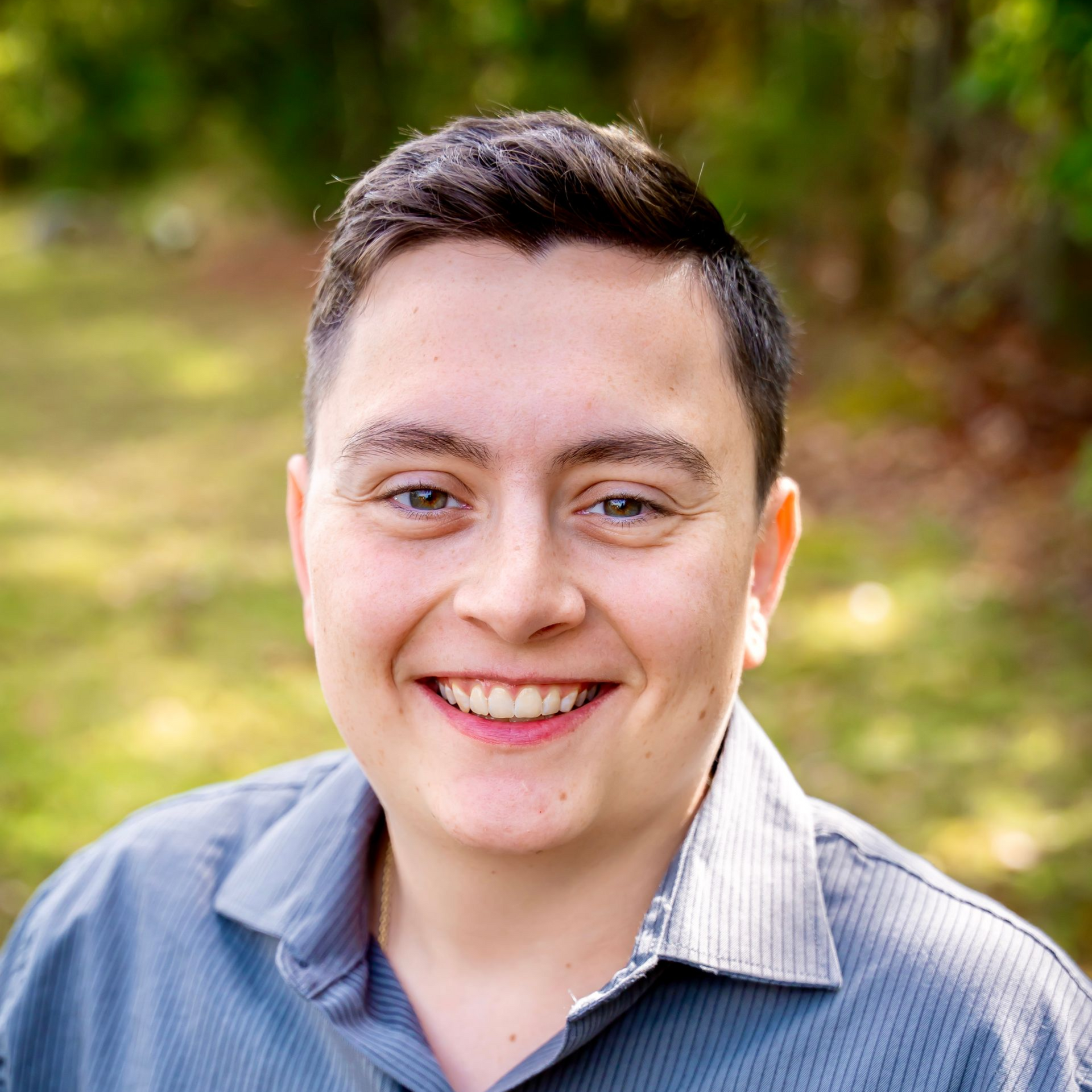 Person smiling, wearing a blue button-up shirt, against a blurred green background.