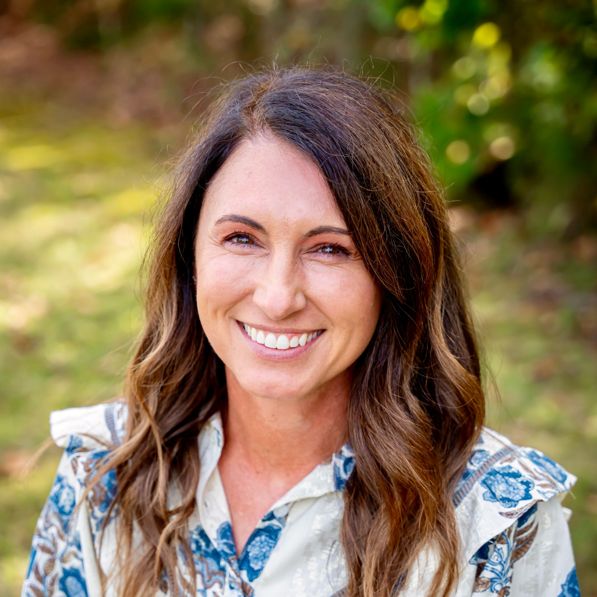 Woman with brown hair smiles, wearing a patterned blue and white dress, sitting outside.