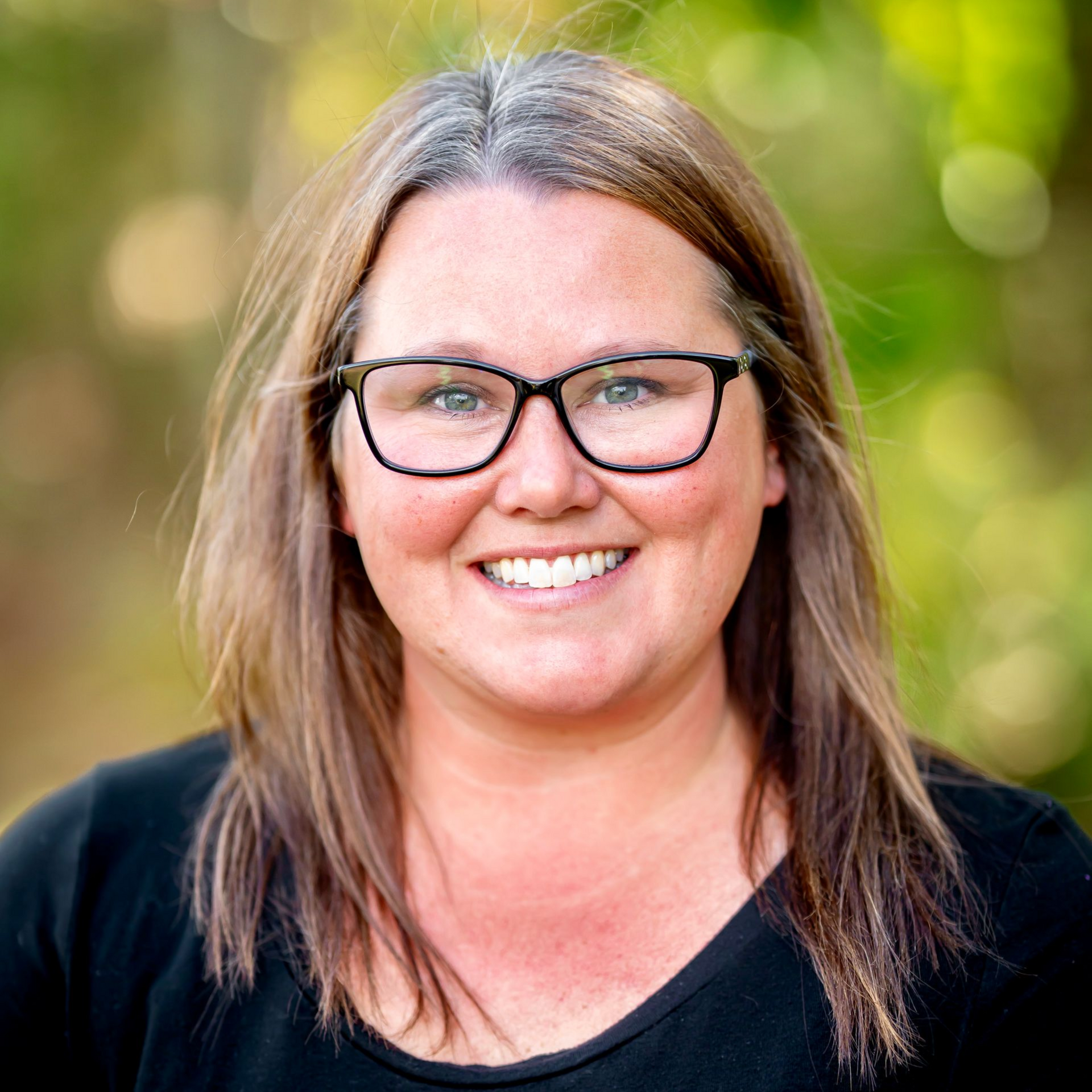 Woman with glasses smiles; outdoors, blurred green backdrop, wearing black top.