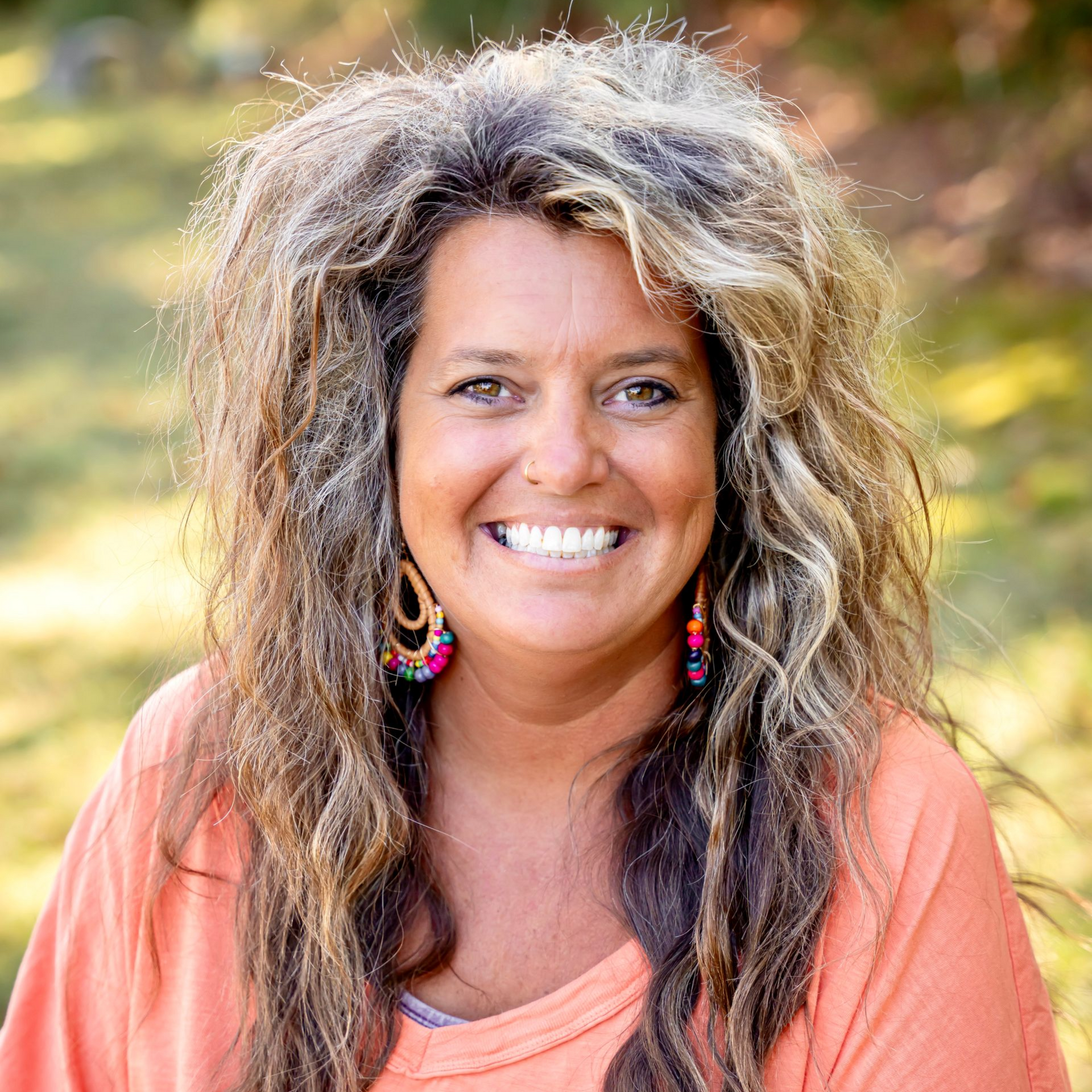 Woman with long curly hair smiles outdoors, wearing an orange top and hoop earrings.