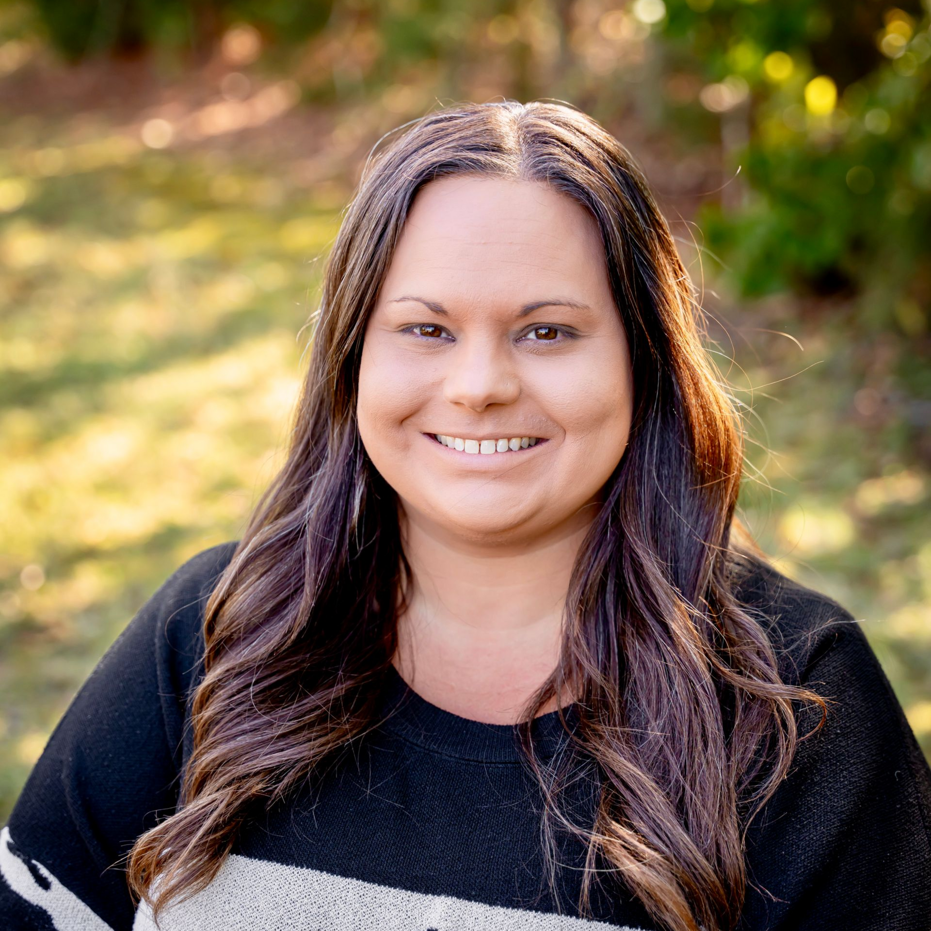 Woman with long brown hair smiles, wearing black and white patterned sweater outdoors.