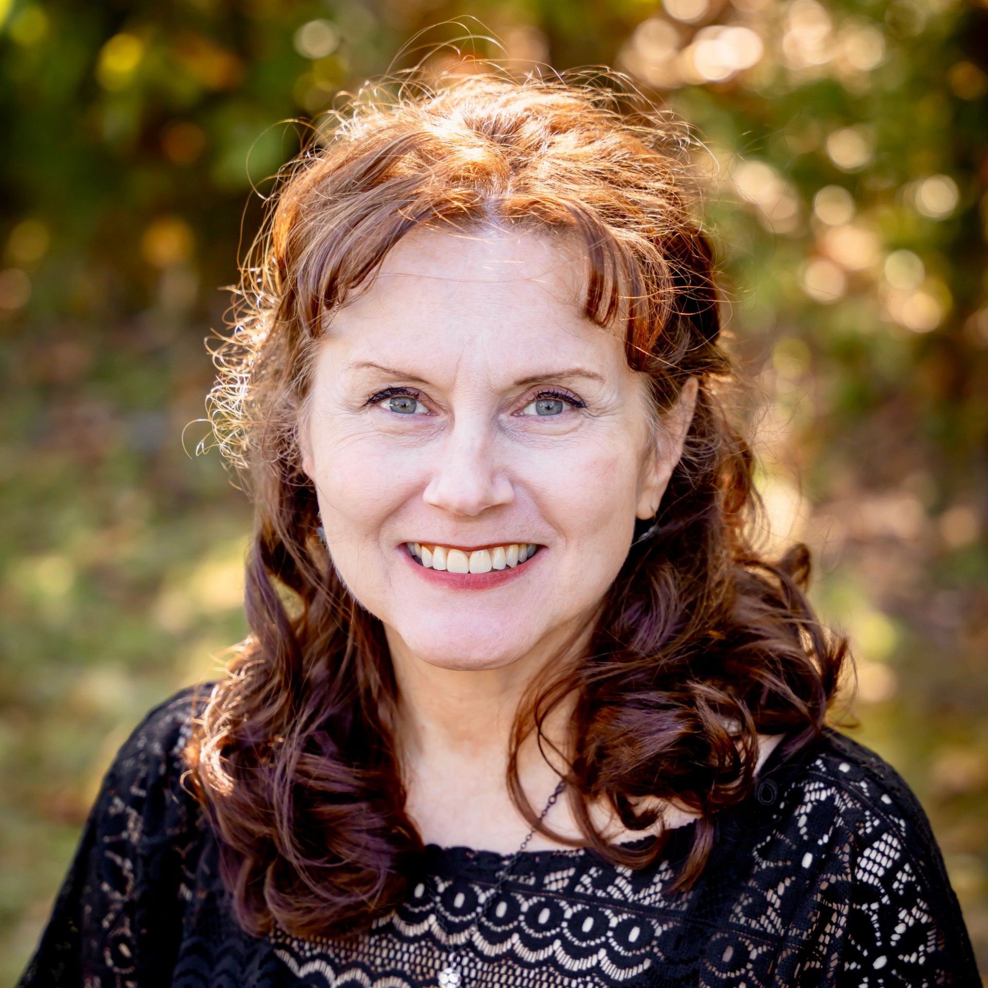 Woman with reddish-brown curly hair smiles at camera, wearing a black lace top outside with blurred foliage.