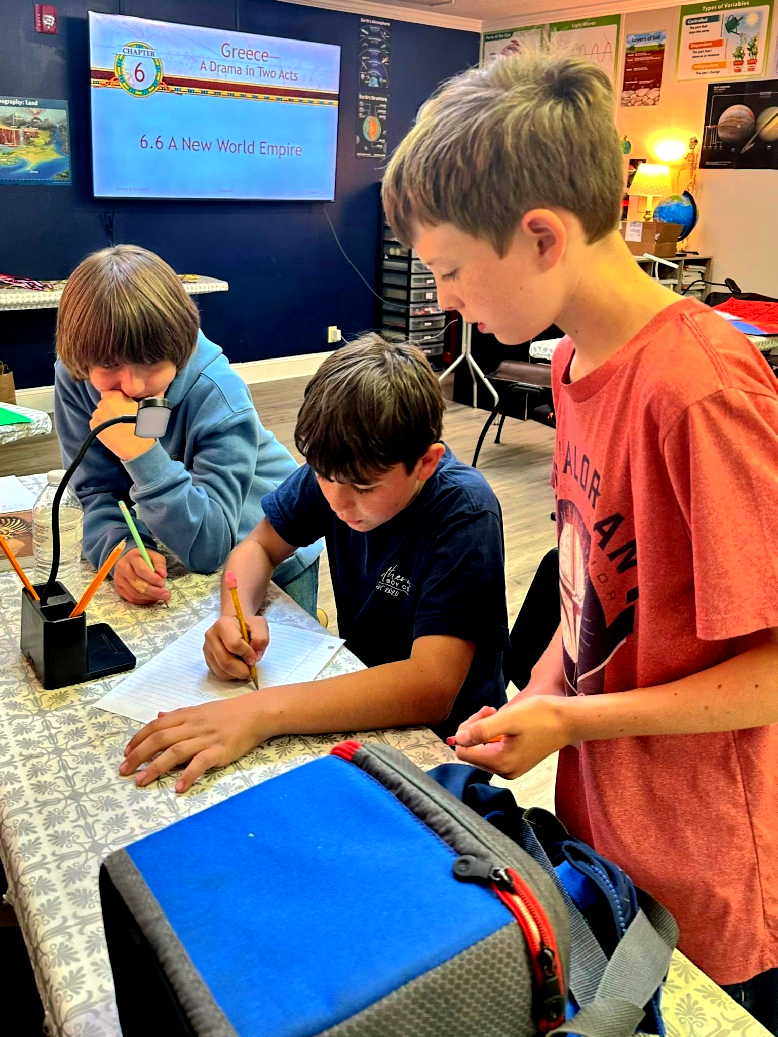 Three boys studying together at a desk. One writes, two watch.