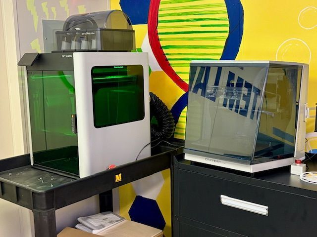 Two 3D printers, one white and one dark, on a cart and a cabinet, against a colorful wall.