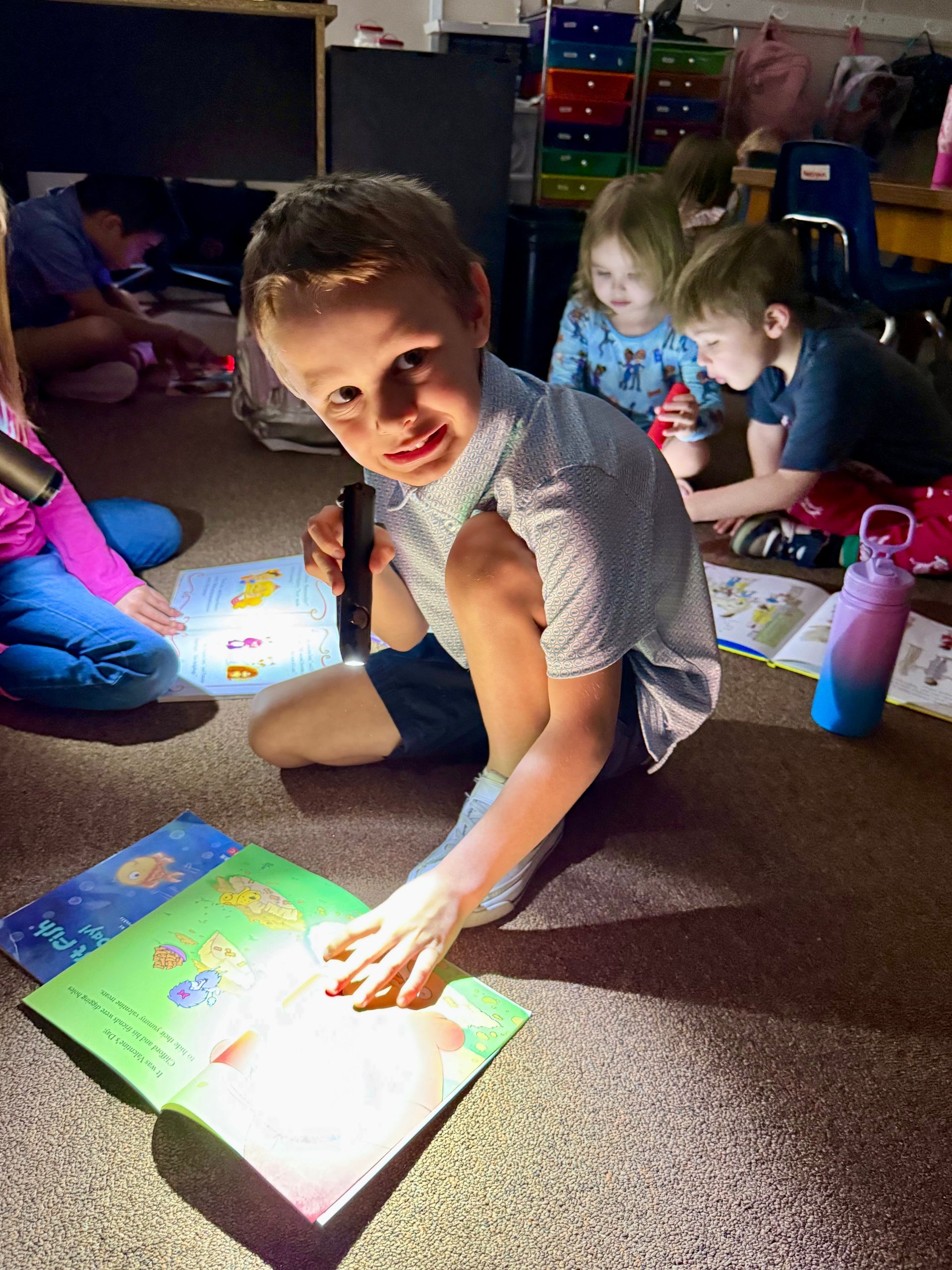 Boy shines a flashlight on a book while sitting on the floor with other children, reading in a dimly lit room.