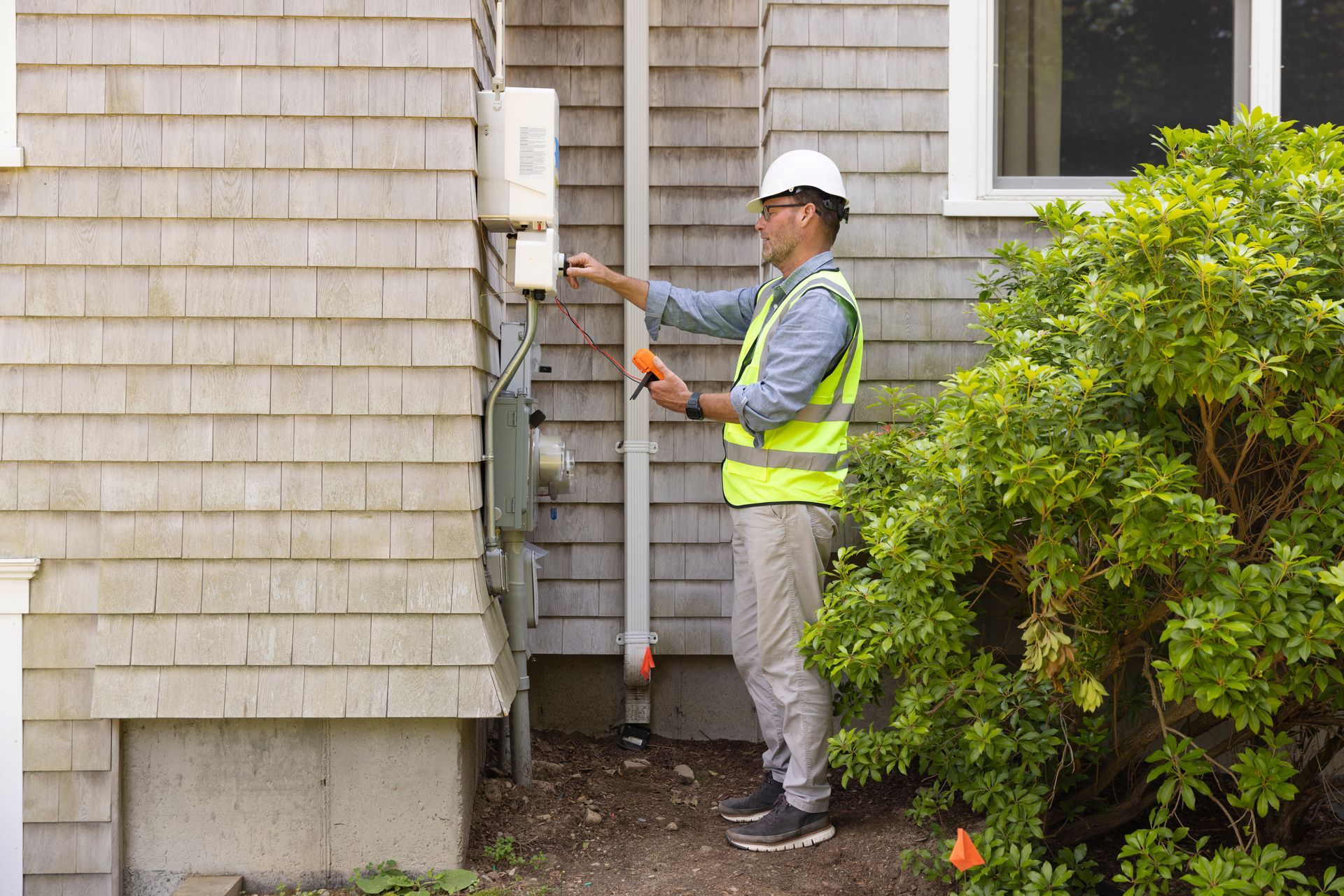 Utility worker inspecting electrical meter beside a house, holding a tool near the siding.