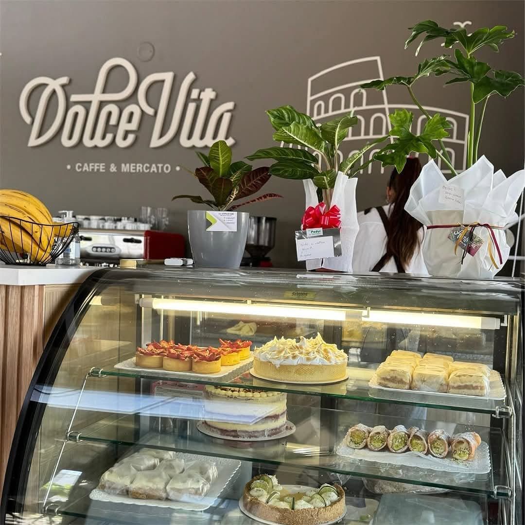 Vitrina de cafetería con pasteles y postres bajo el letrero de Dolce Vita y plantas en macetas.