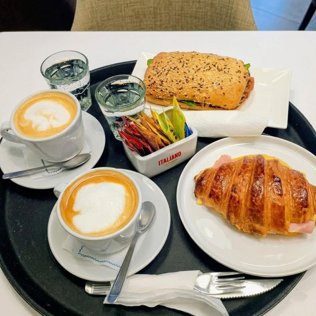 Café y bollería en una bandeja: dos lattes, un cruasán, un pan de semillas y sobres de azúcar.