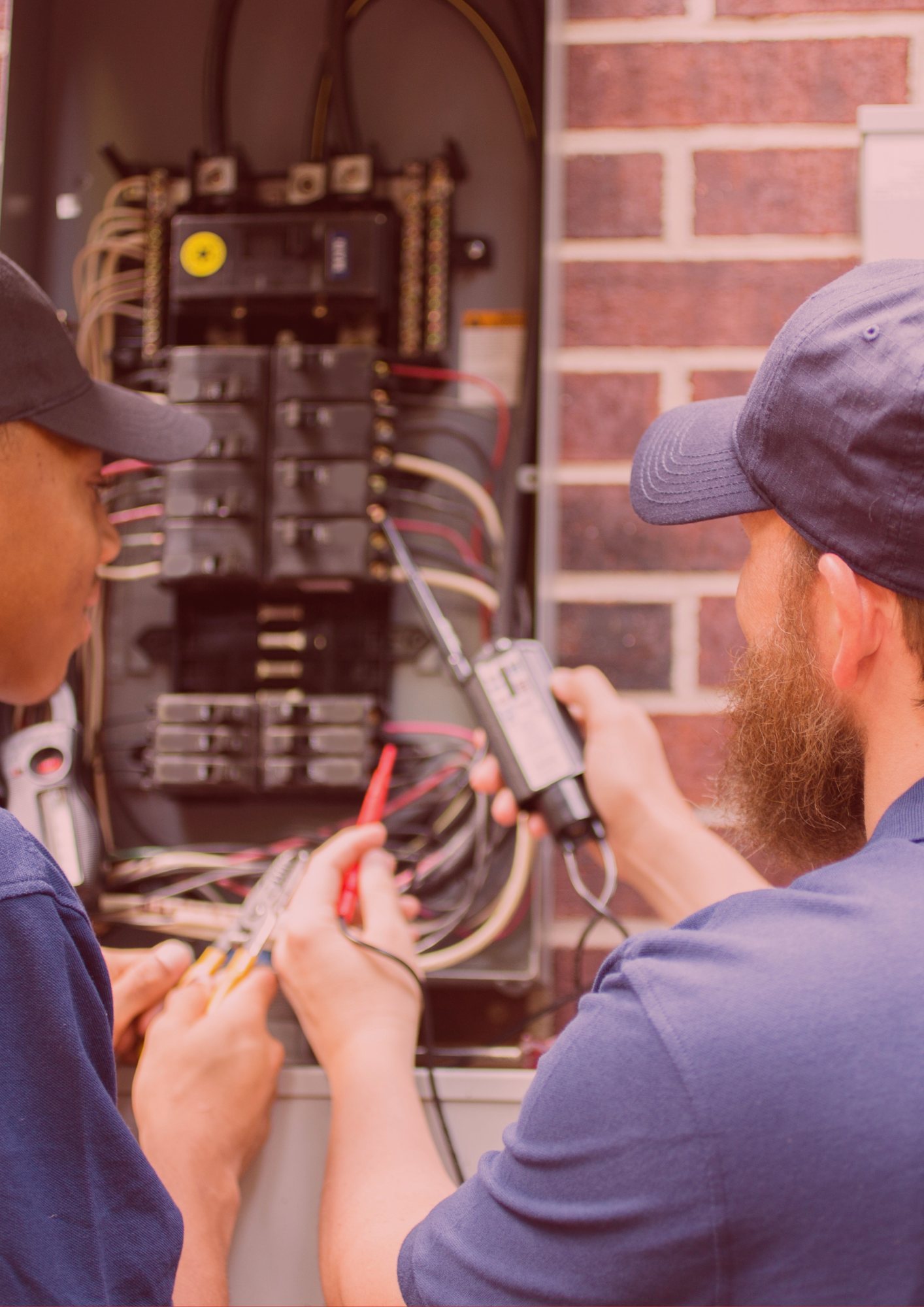 Two men are working on a electrical box in front of a brick wall.