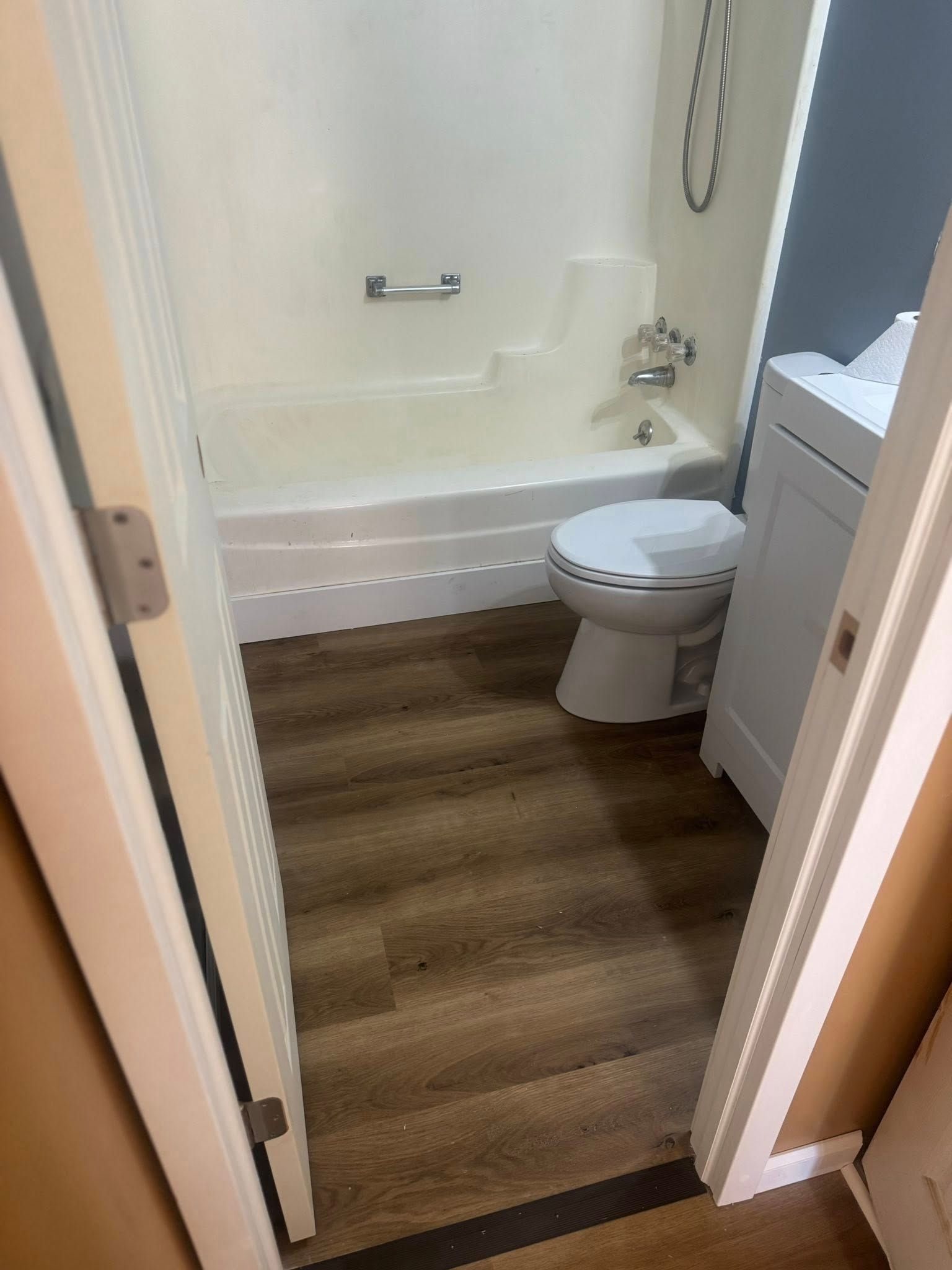 Small bathroom with white tub, toilet, vanity, and wood-look floor seen through an open doorway.