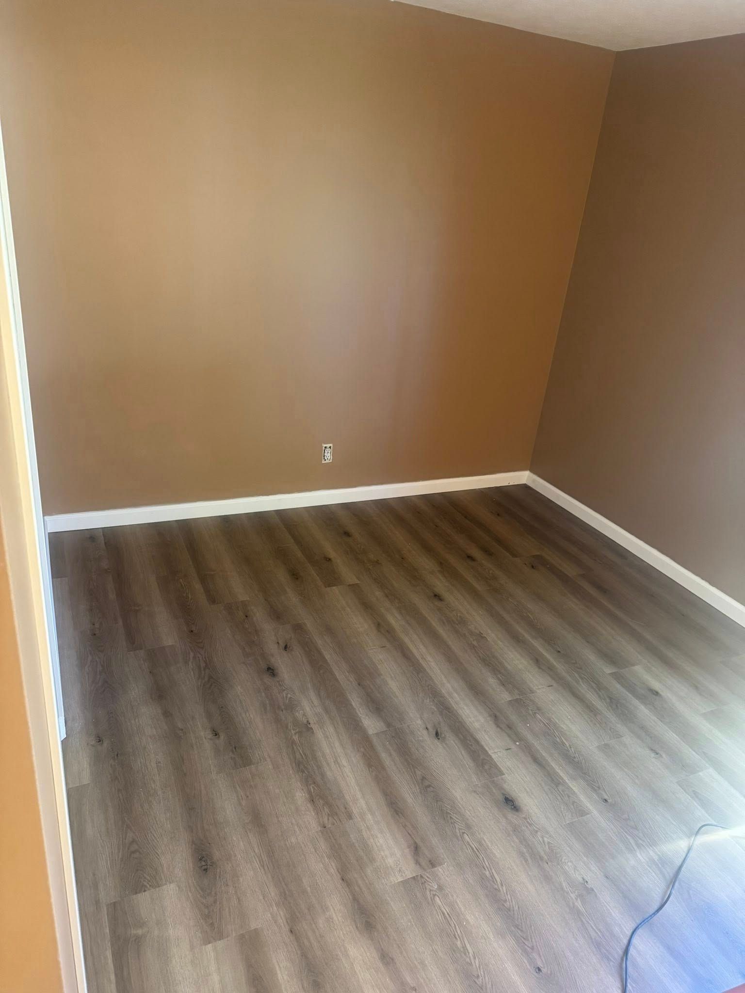 Empty room with brown walls, wood flooring, and white baseboards.