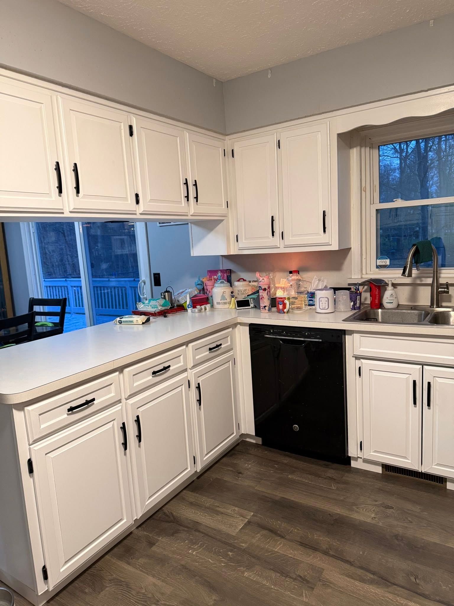 White kitchen with cabinets, countertops, black dishwasher, wood floor, and two windows looking outside.