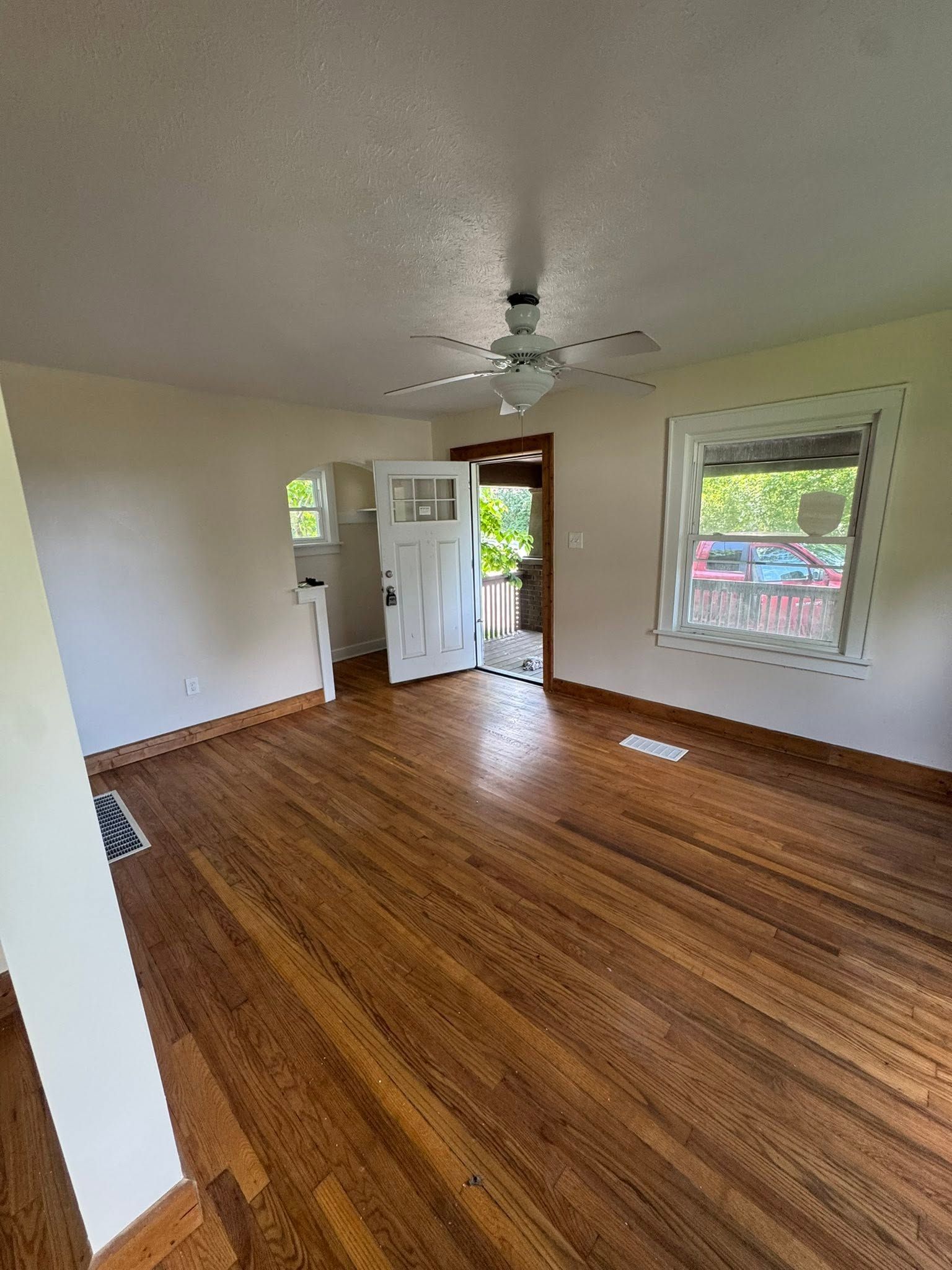 Empty room with hardwood floors, white walls, ceiling fan, and sunlight through windows