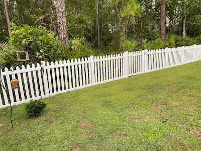 White picket fence in a grassy yard with trees in the background.