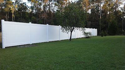 White vinyl fence in a grassy yard with a tree, with a treeline in the background.
