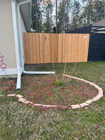 A wooden fence and a curved flower bed bordered by bricks sit near a house's corner and a black fence in a backyard.
