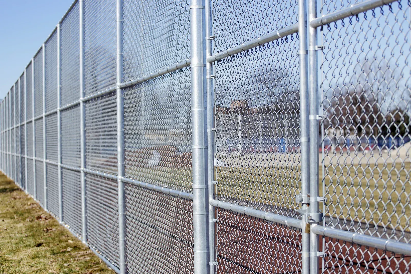 A chain-link gate with a silver latch, attached to gray metal posts, with a tennis court visible in the background.