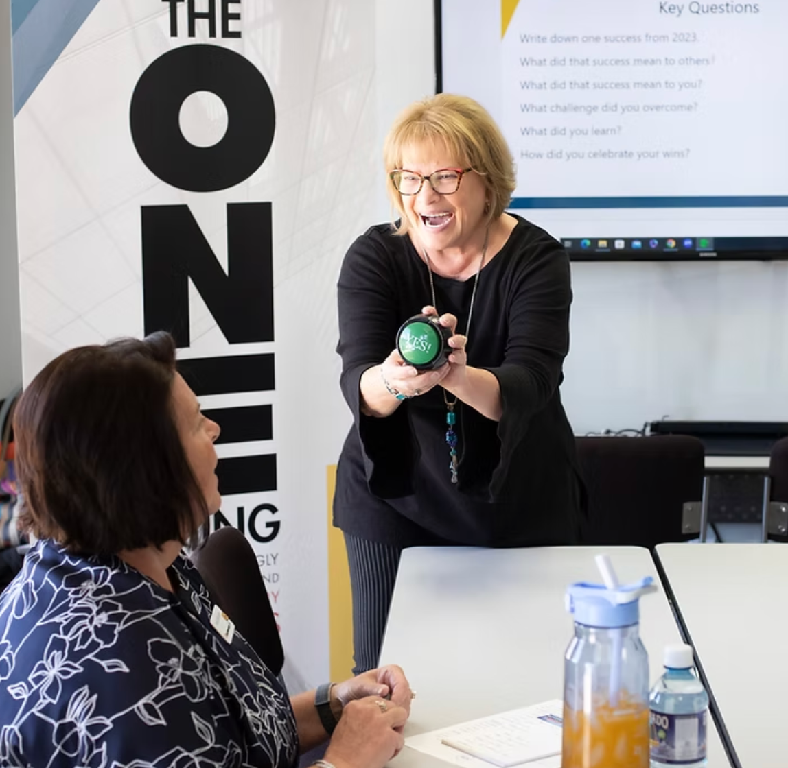 Woman smiling and holding a green device during a classroom demonstration with seated participants.