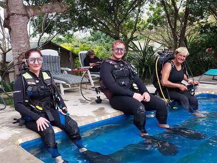 Three scuba divers in black wetsuits sit at a pool edge, smiling and resting with gear on.